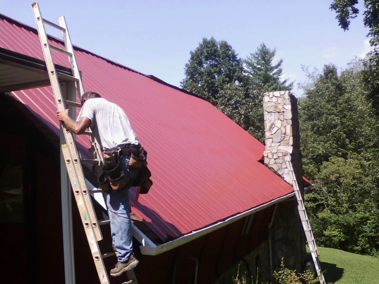 Roofer on a ladder beside a red metal roof and stone chimney — Soffit & Fascia Repair by Roofing Brothers in Leigh